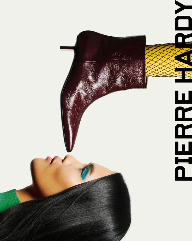Fashion editorial featuring a burgundy ankle boot hovering above a woman's face with teal eye makeup against a stark white background.