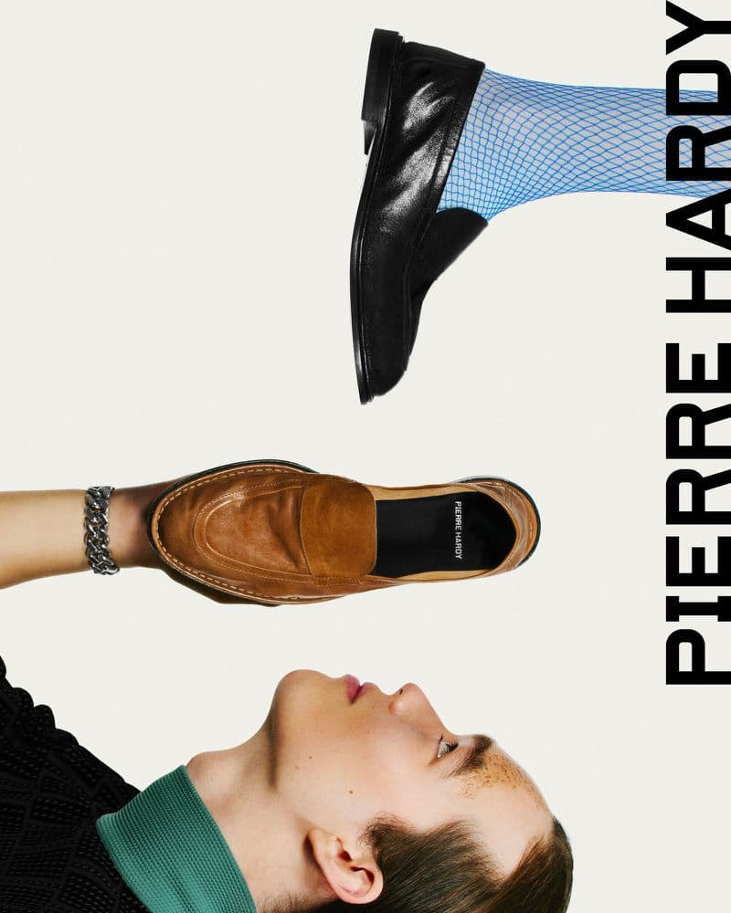 Fashion editorial shot featuring a man's face and two loafers, one brown held by a hand, and the other black on a blue fishnet-clad foot.