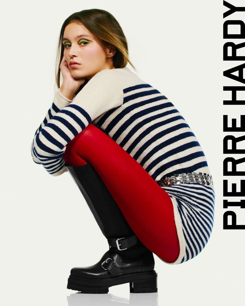 Fashion shot of a young female model crouching in a striped sweater dress, red tights, and black boots against a white backdrop.