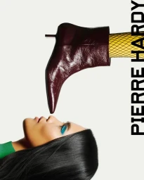 Close-up portrait of a woman with turquoise eye makeup and a burgundy leather boot pointed near her nose against a plain white background