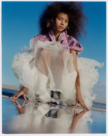 Fashion shot of a young woman with dark curly hair, wearing a white dress, crouching against a clear blue sky with reflection.