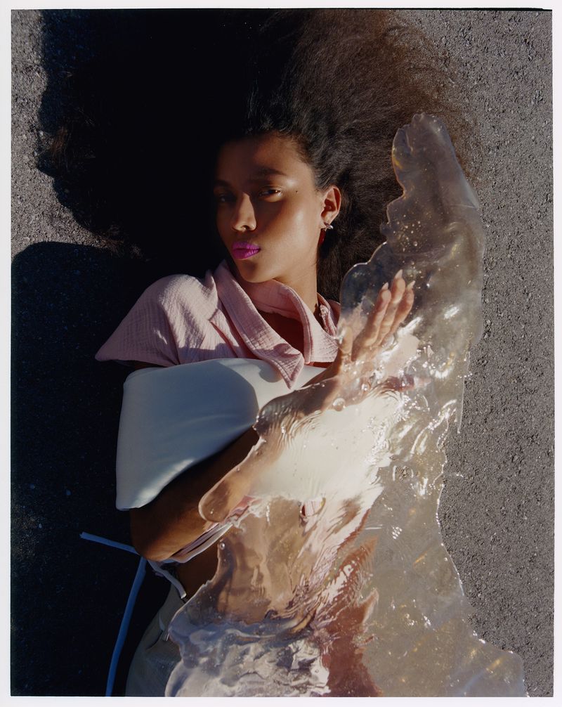A woman with dark curly hair lies on asphalt, holding a translucent object, eyes closed, wearing a pink top and light-colored pants.