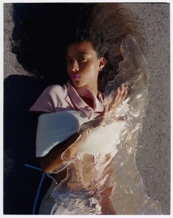 A woman with dark curly hair lies on asphalt, holding a translucent object, eyes closed, wearing a pink top and light-colored pants.