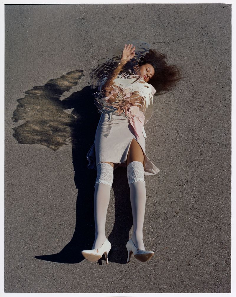 A woman with dark hair laying on grey asphalt in a beige mini skirt, white top, white stockings, and heels with a surreal shadow.