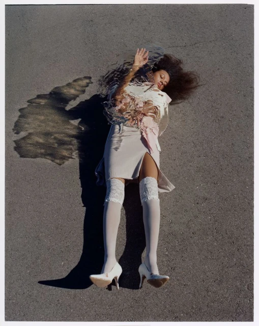 A woman with dark hair laying on grey asphalt in a beige mini skirt, white top, white stockings, and heels with a surreal shadow.
