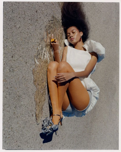 An adult female lies on a concrete surface, wearing a white dress with ruffles, orange stockings, and floral heels, holding a yellow lighter.