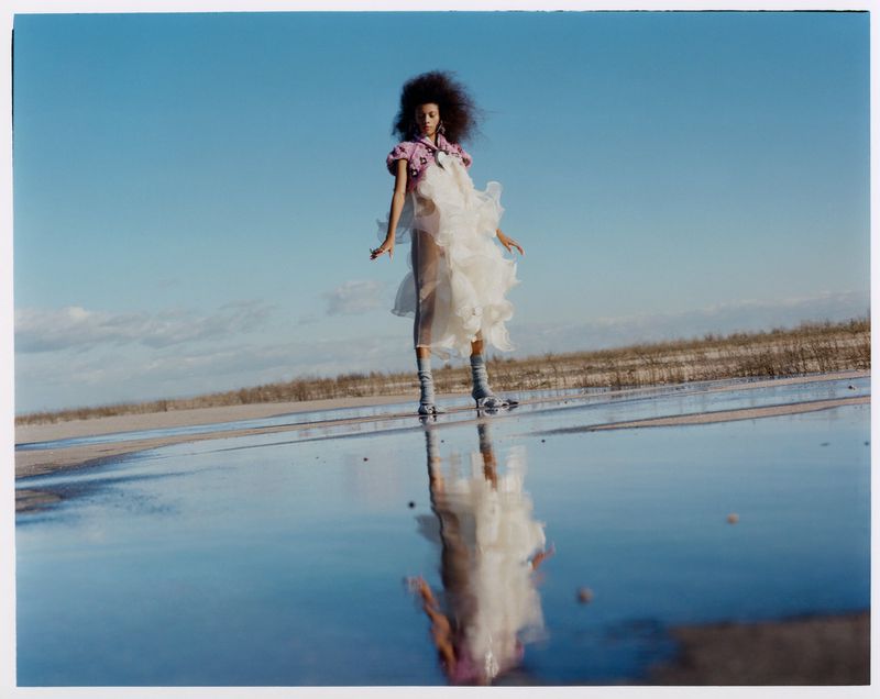 A woman with an afro poses in a white dress and purple top on a reflective salt flat under a blue sky, creating a mirrored effect.