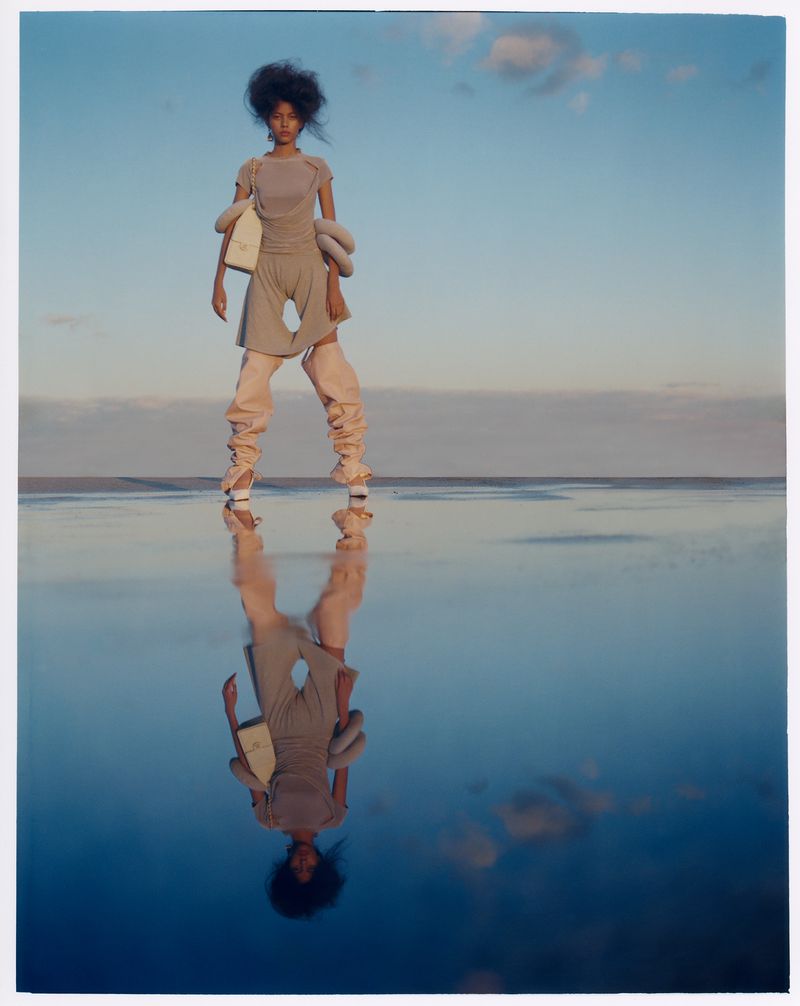Woman with afro poses in beige jumpsuit with oversized sleeves and leg warmers, reflected on a water surface with a blue sky.