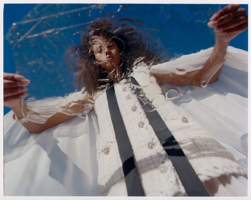 Woman in a white dress poses with a transparent material against a clear blue sky, looking up towards the camera, low angle shot.