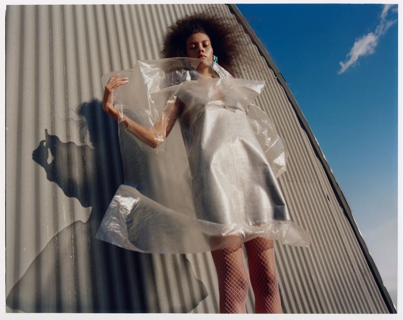 Young woman with curly hair lies on a silver metallic surface outdoors, wearing a transparent plastic dress and fishnet stockings.
