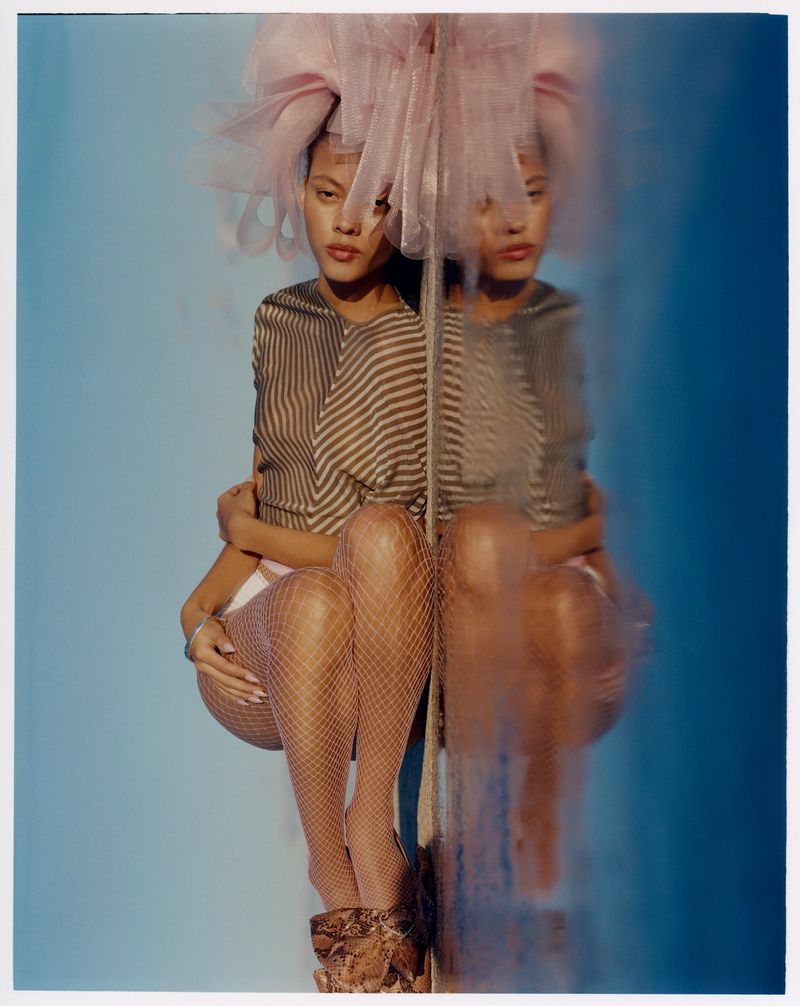 A woman with brown hair poses with reflection. She wears brown and tan striped top, fishnet stockings, and high heels against blue backdrop.