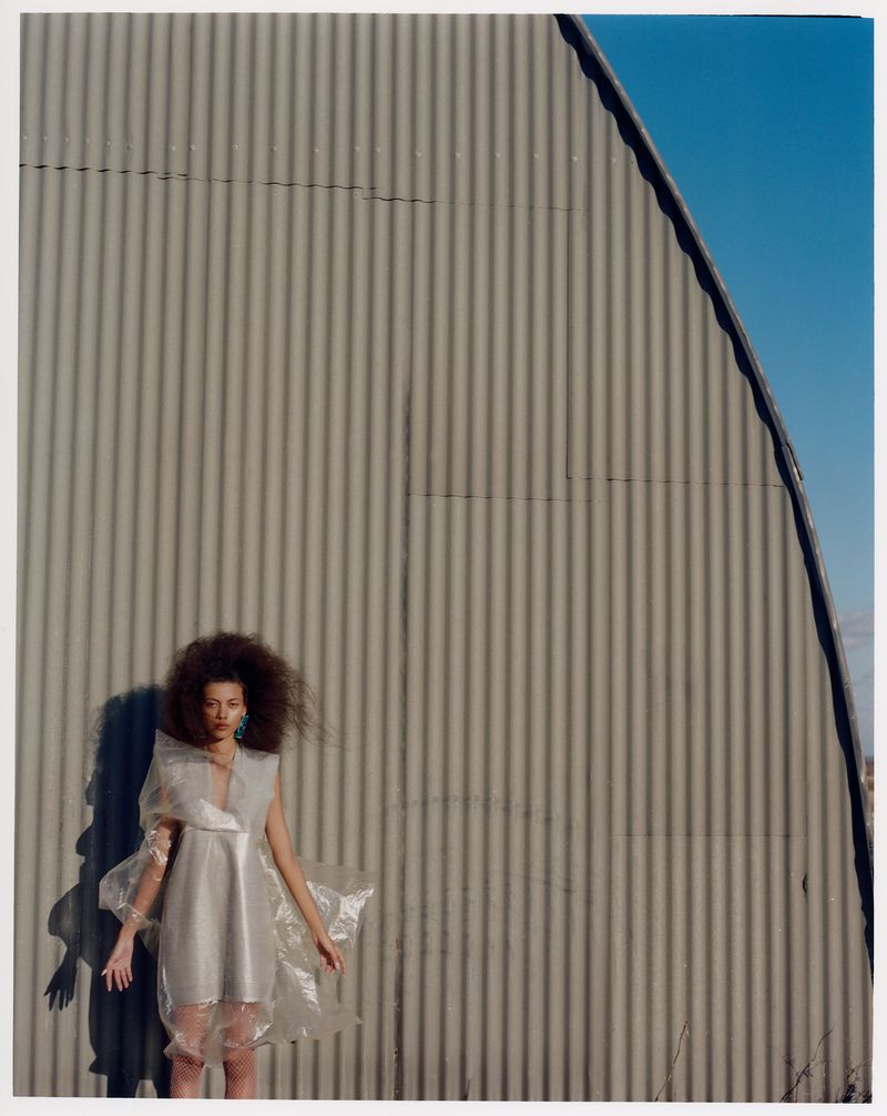 Fashion shot of a woman with afro posing in a transparent dress against a corrugated metal building under a bright blue sky.