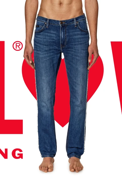 Torso shot of a person wearing denim jeans, standing against a backdrop featuring the logo and the word LOVE, with bare feet.