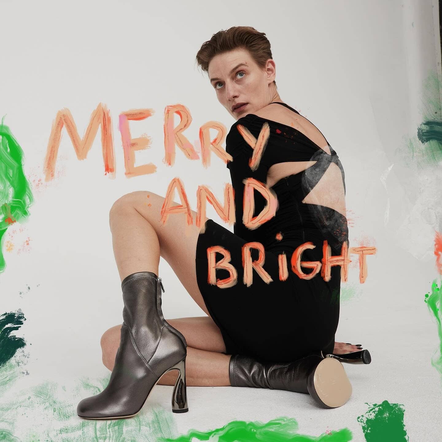 Woman poses seated in a short black dress and metallic ankle boots against a painted white backdrop with holiday text.