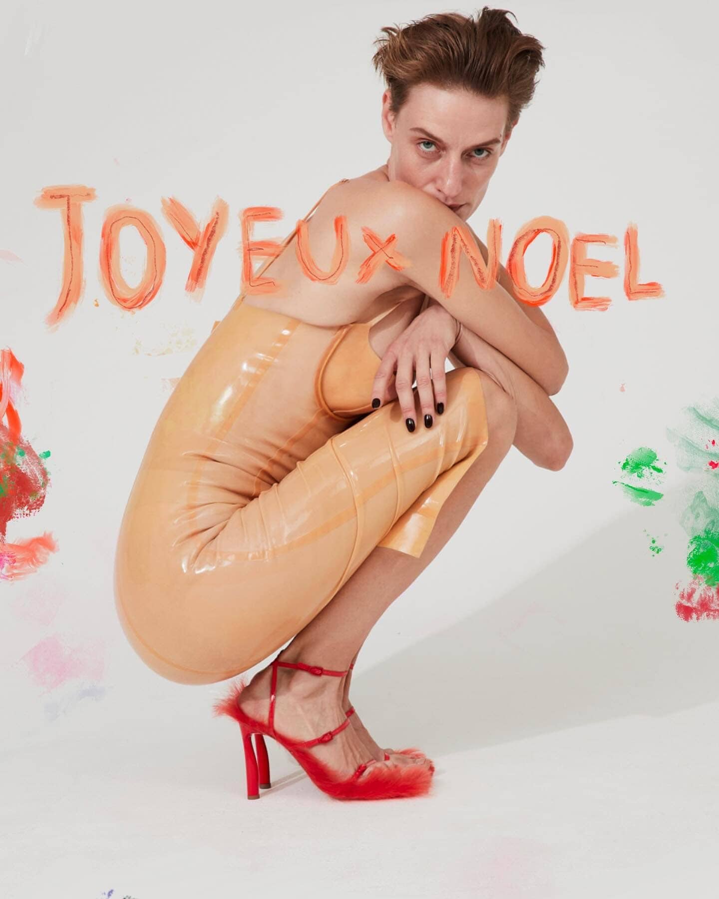 Woman in tan latex dress and red heels crouching in a white studio with red and green art and the phrase "Joyeux Noel".