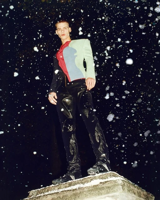 Fashionable adult male model in colorful jacket and black leather pants stands on concrete platform against a dark, splattered background.