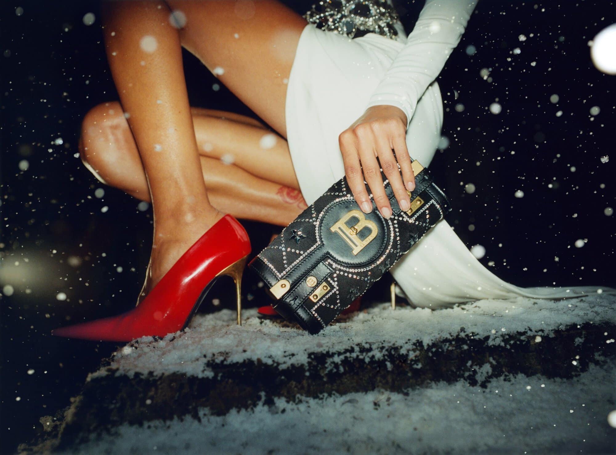 A handbag and red heels are featured in a fashion shot with snow. The model is posed in a white dress against a dark backdrop.