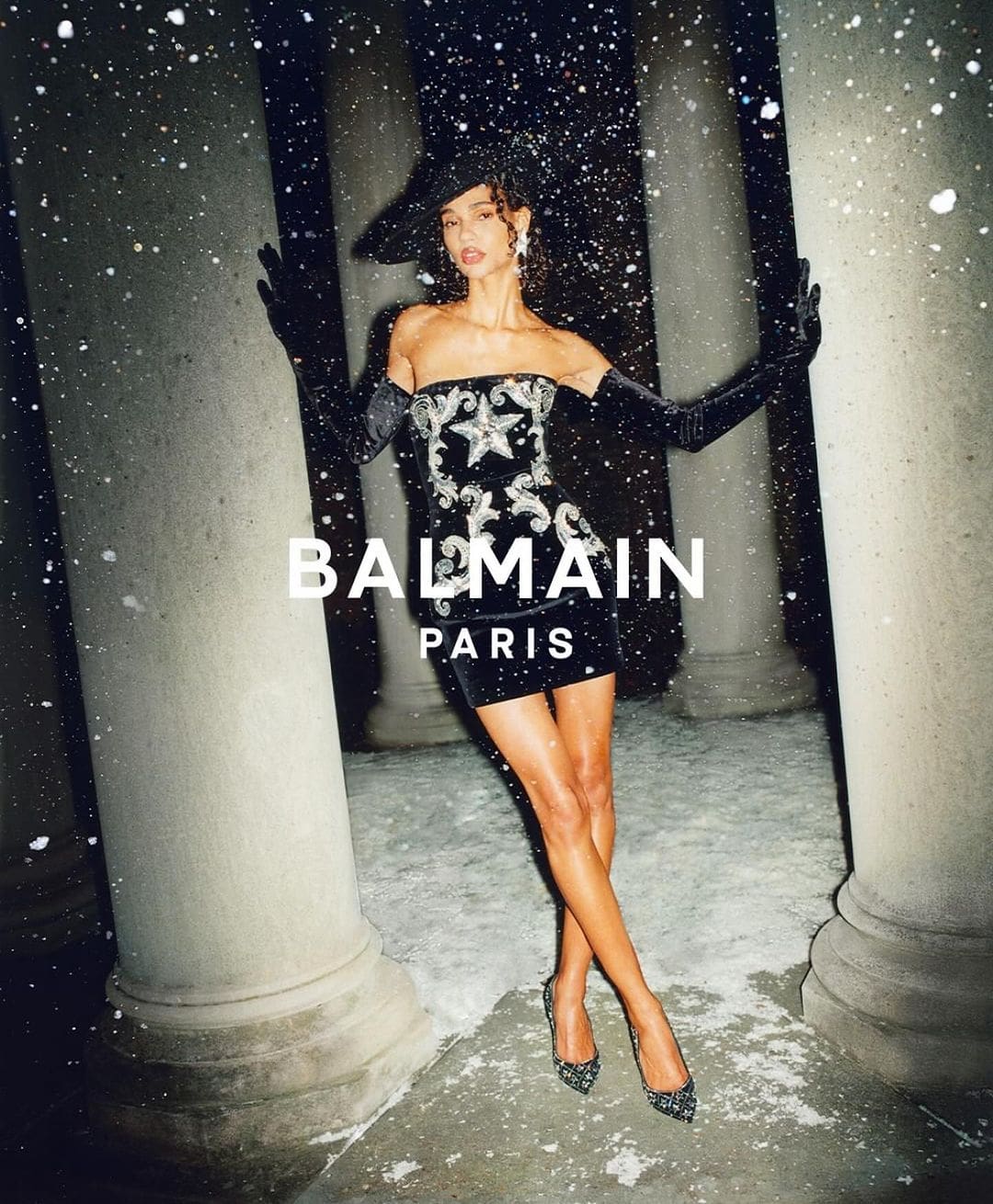 Fashion shot of a woman in a black dress and black gloves, posing near stone pillars with snow falling. Balmain Paris logo visible.