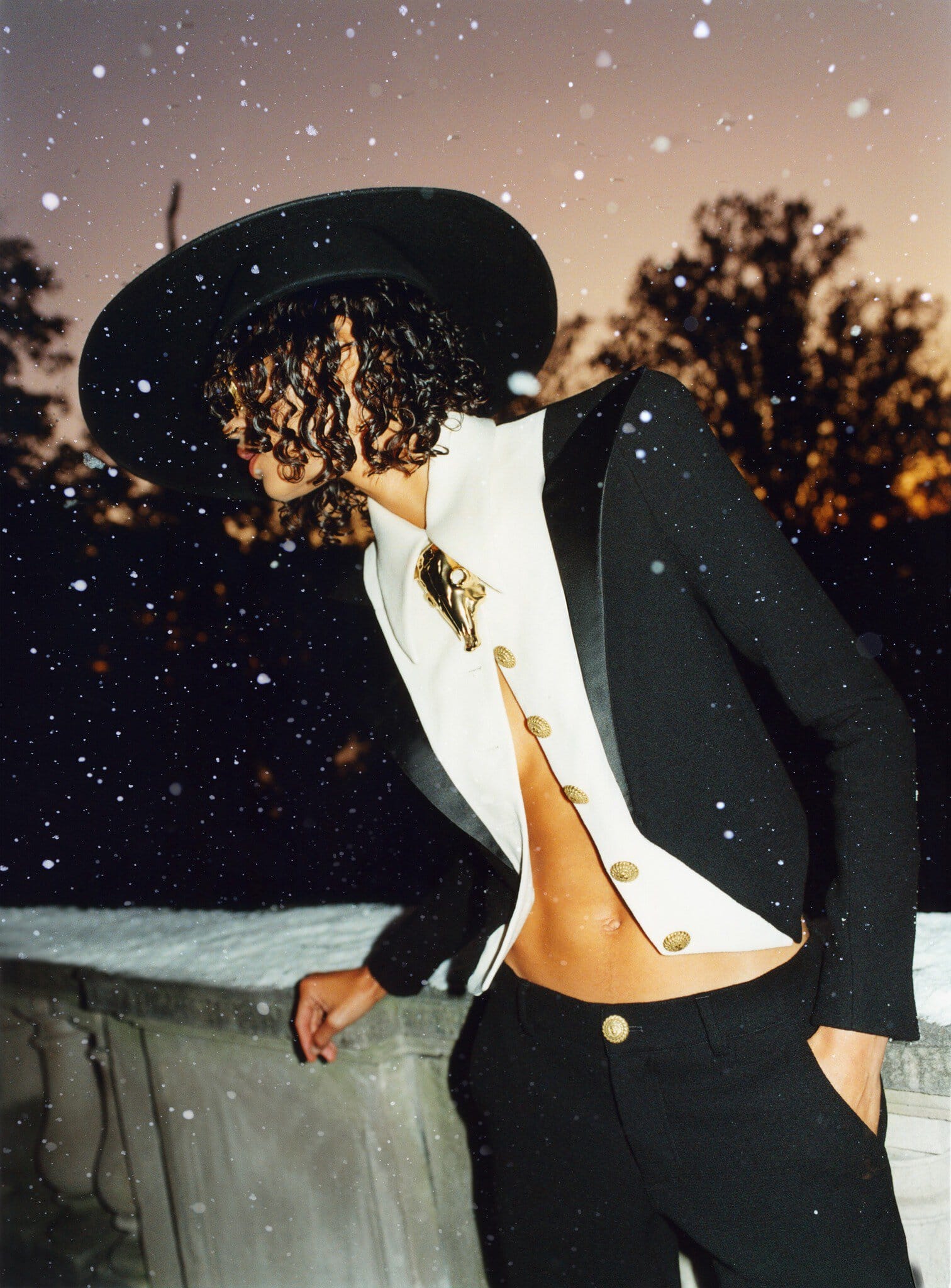 Fashion shot of a person in a black hat and jacket, and white dress shirt with gold buttons, posing on a stone balcony outdoors.