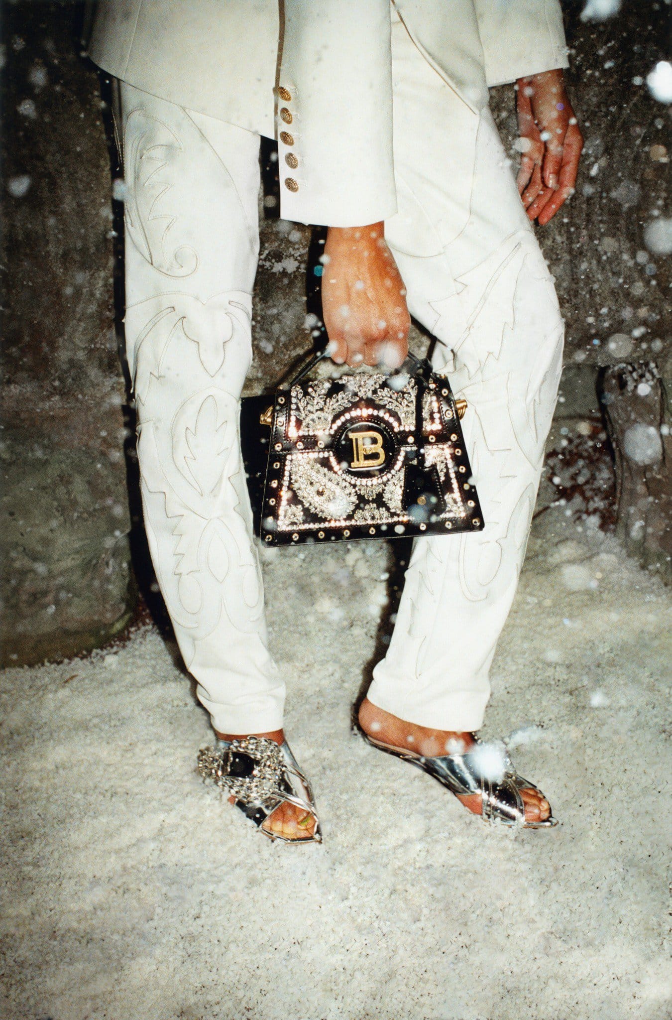 Fashionable person stands in snow wearing a white suit, embellished sandals, and holding an embellished black Balmain purse.