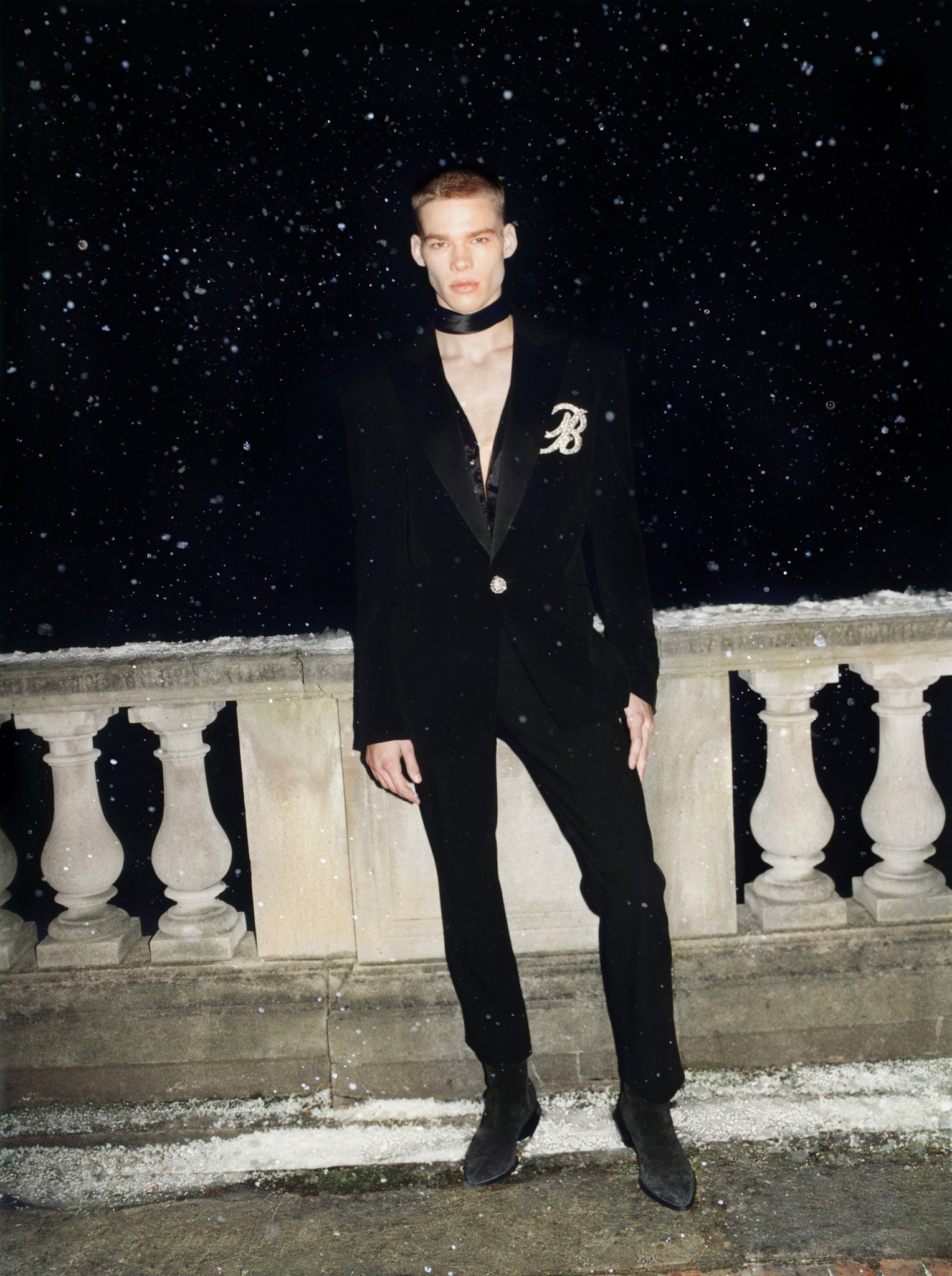 Male fashion model poses outdoors at night in a black suit with a black ribbon and jewel embellishment during a light snowfall.
