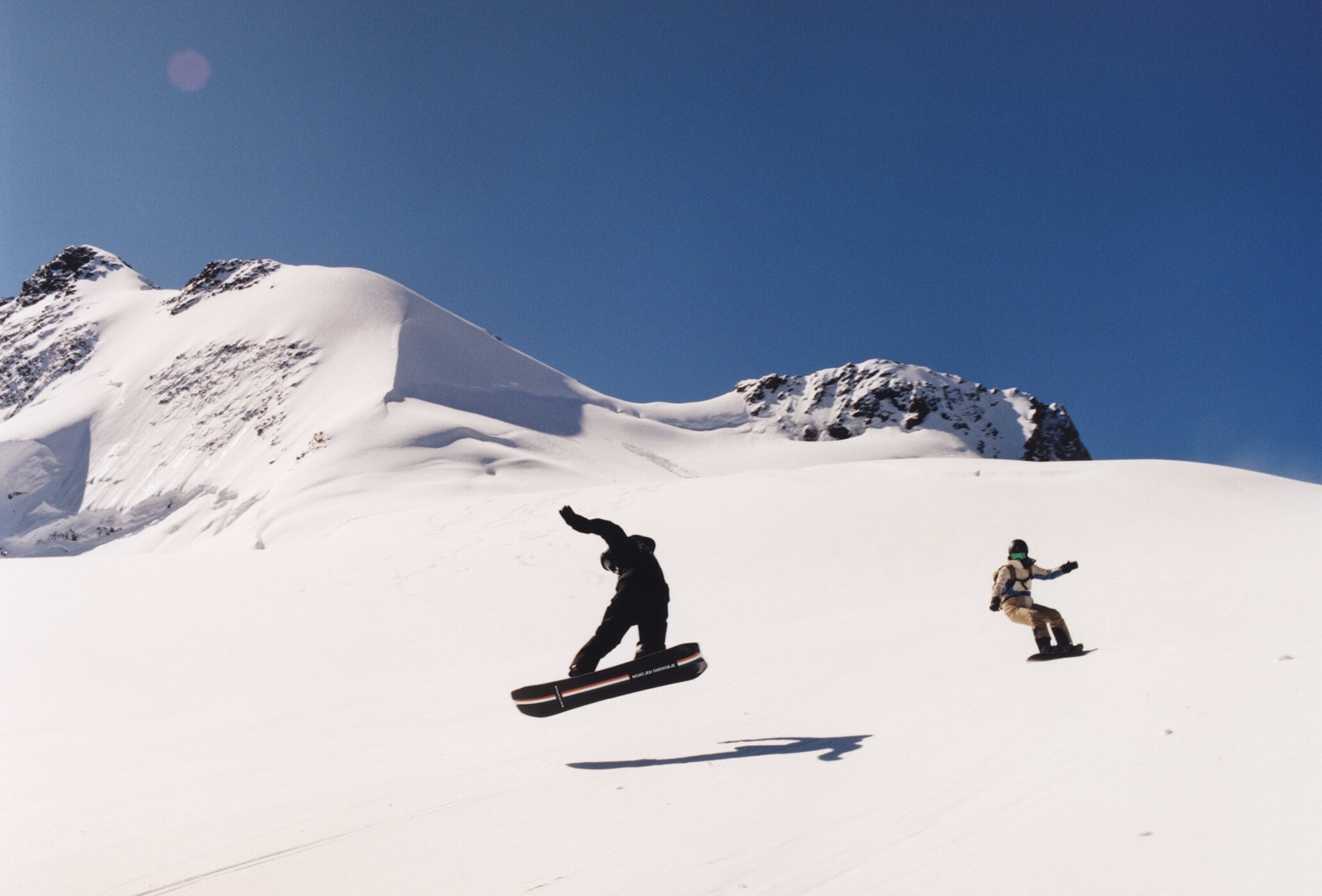 Two snowboarders are airborne against a snowy mountain backdrop and blue sky, showcasing winter sports in a vast outdoor setting.