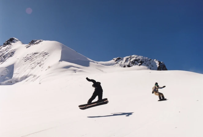 Two snowboarders are airborne against a snowy mountain backdrop and blue sky, showcasing winter sports in a vast outdoor setting.