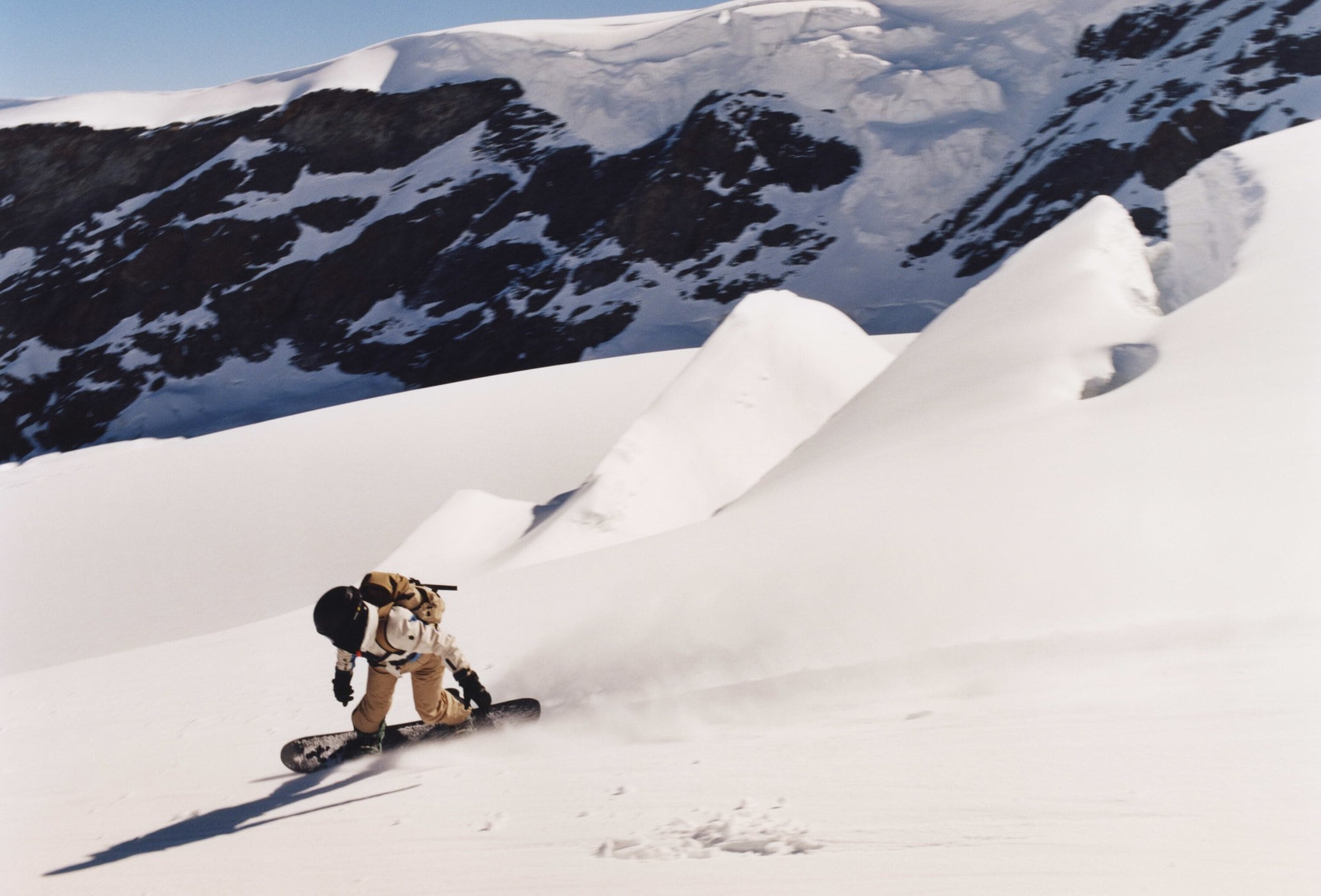 Snowboarder rides down a mountain slope with snow-capped mountains in the background on a sunny day, creating a dynamic winter scene.