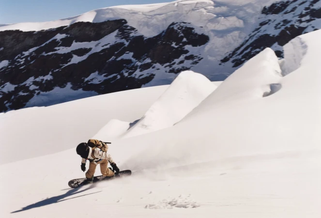 Snowboarder rides down a mountain slope with snow-capped mountains in the background on a sunny day, creating a dynamic winter scene.