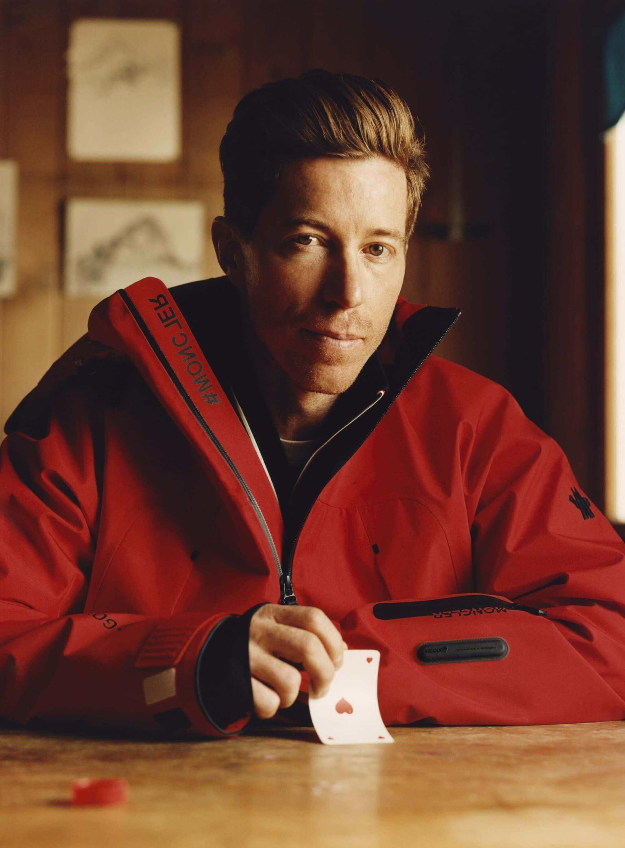 Close-up of snowboarder Shaun White in a red Moncler jacket holding a playing card with a heart suit, set against a rustic backdrop.