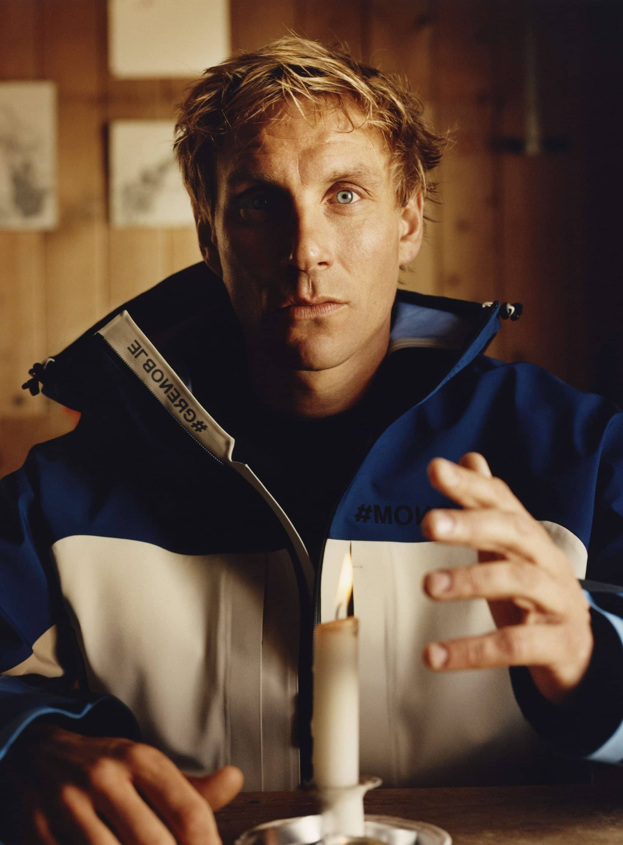 Man in a blue and white jacket sits at a wooden table with a lit candle in a silver holder; indoor setting with wood paneling.