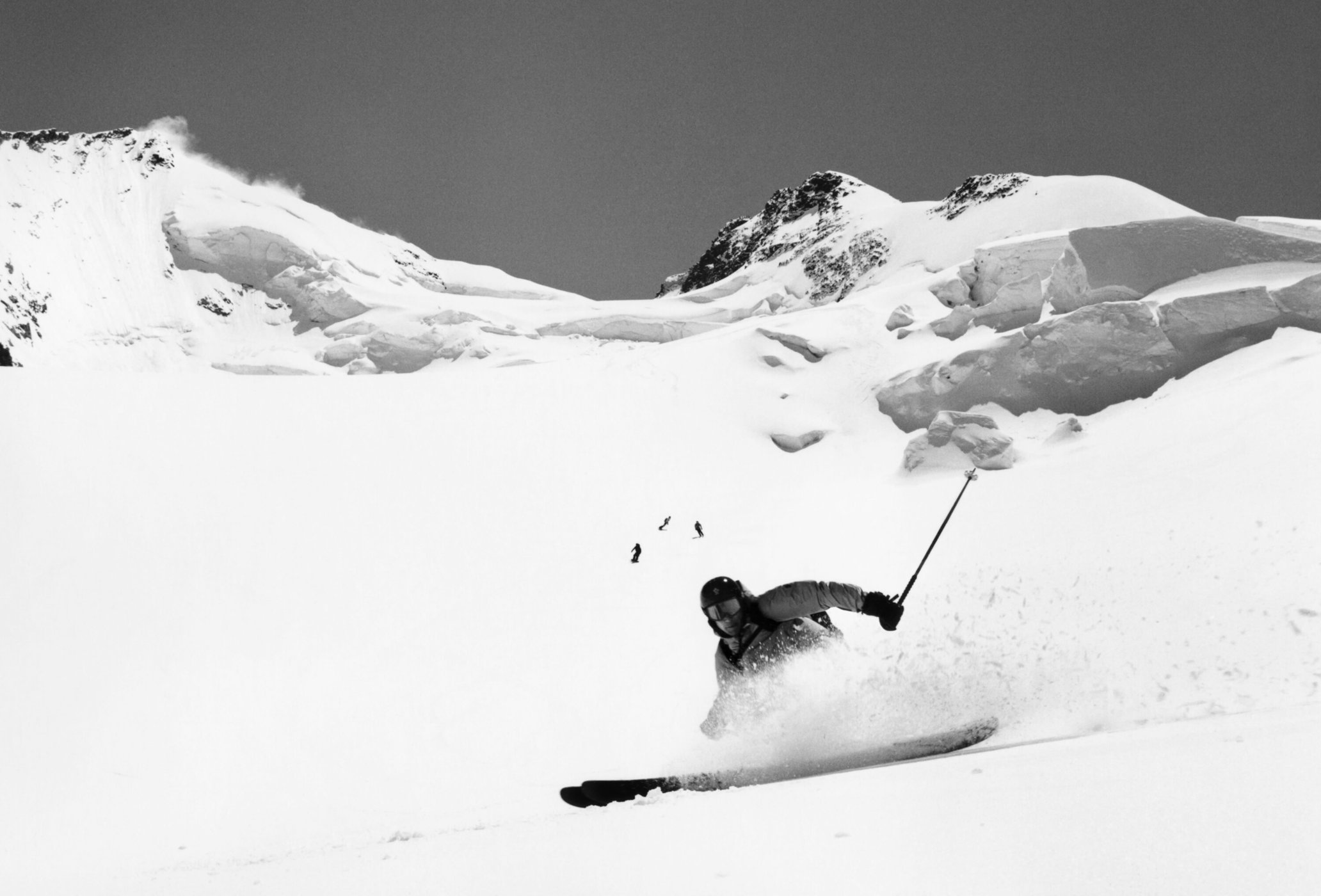 Monochrome action shot of a skier gliding down a snow-covered mountain, with other skiers in the distance and a majestic mountain backdrop.