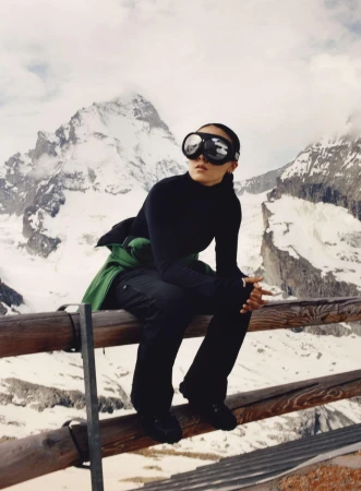 Woman in ski goggles sits on wooden fence against snow-covered mountain backdrop, wearing black turtleneck, dark pants and jacket.