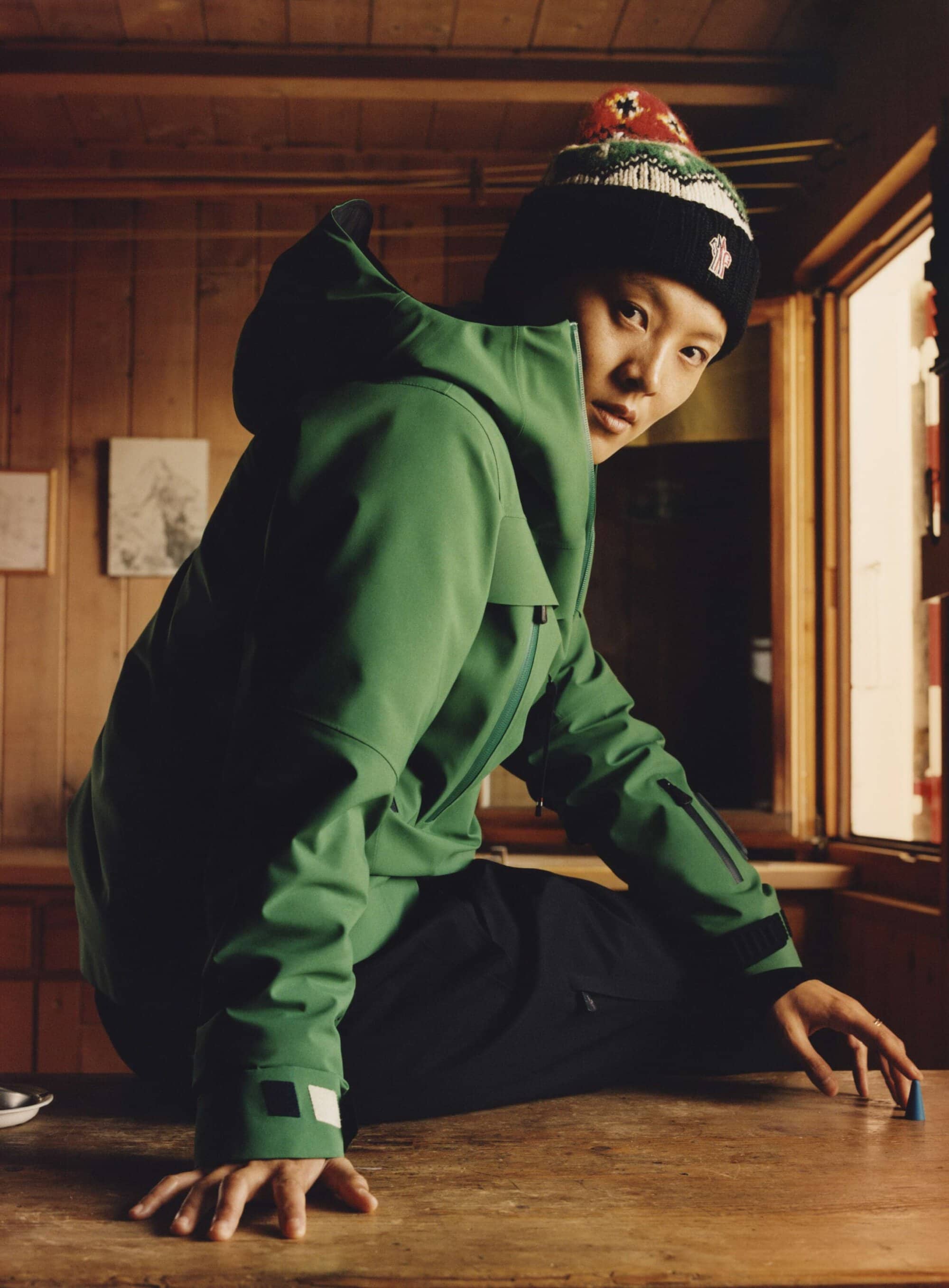 Asian adult wearing green jacket and beanie poses indoors near window. Jacket and hat are primary subjects. Background is neutral.
