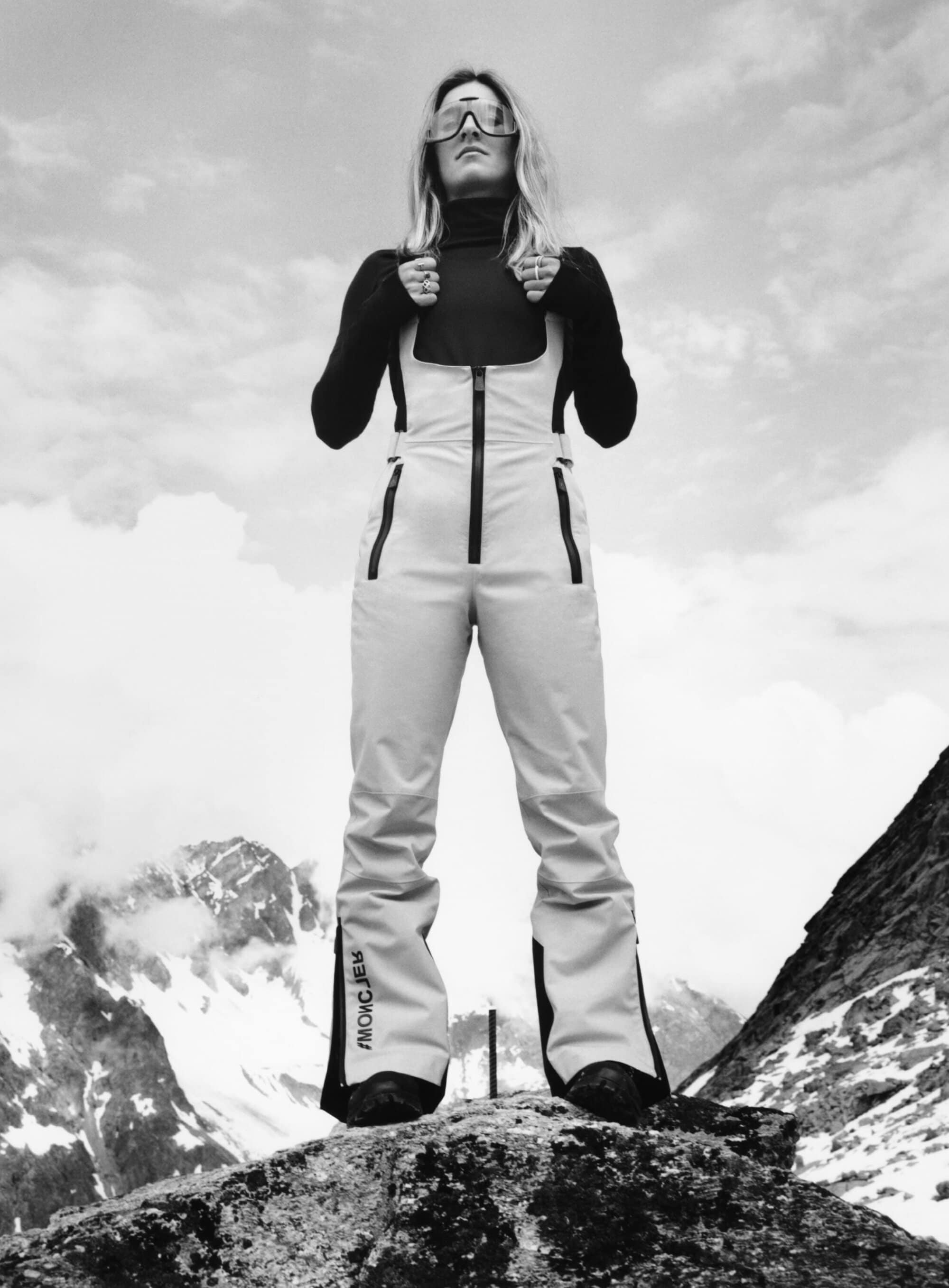 Monochrome portrait of a confident woman in ski goggles and overalls standing on a rock with mountains and clouds in the background.