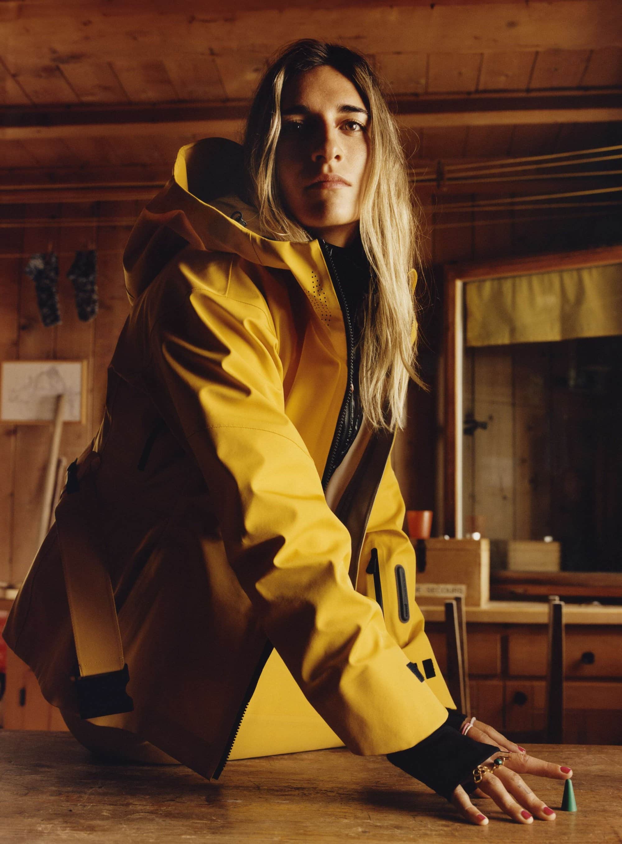Woman in yellow raincoat posing indoors with natural lighting, showcasing fashion and style in a rustic cabin environment.