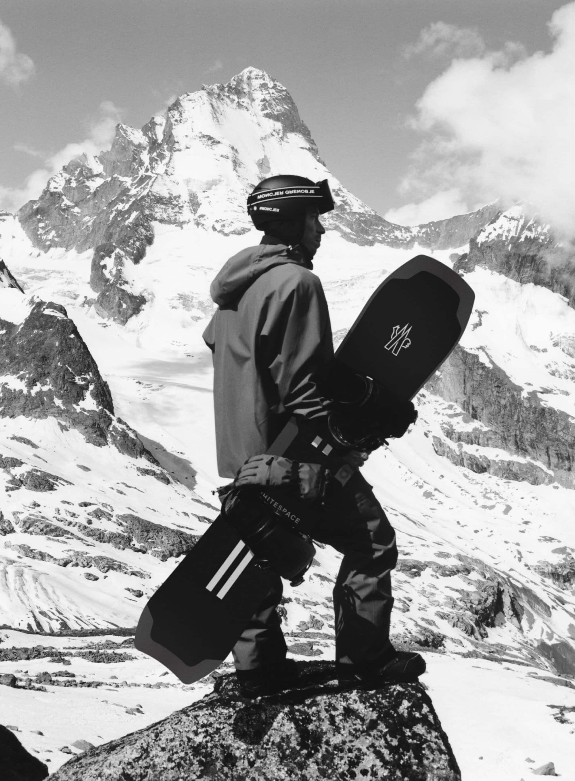 Man with snowboard stands on a rocky peak against a background of snowy mountains and cloudy sky in a black and white outdoor shot.