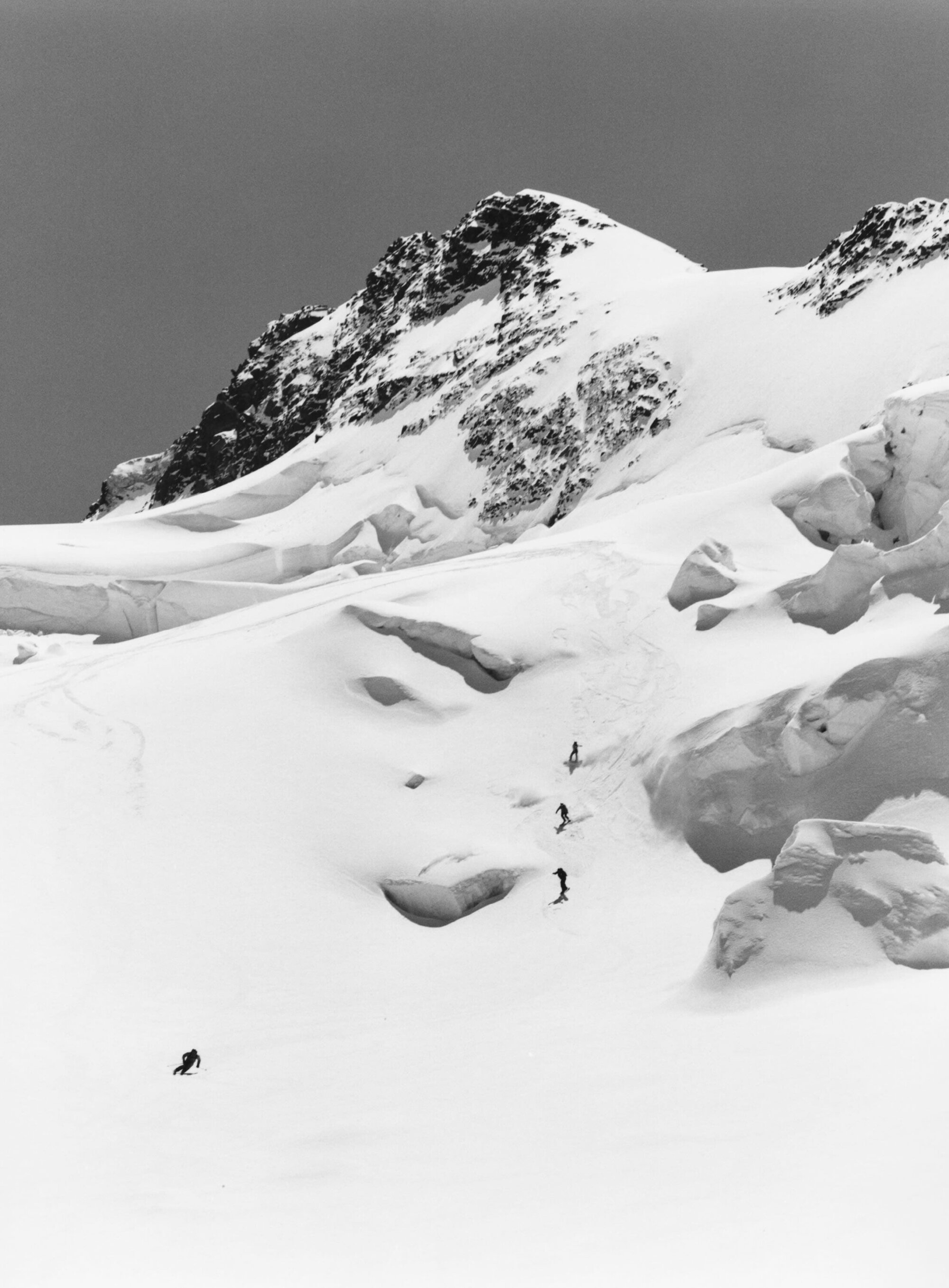 Monochromatic wide shot of a snowy mountain with a group of five people skiing down the slope in the distance, showcasing vastness and isolation.