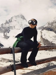 Person wearing black ski goggles and dark apparel seated on a wooden railing against a snowy mountain backdrop.