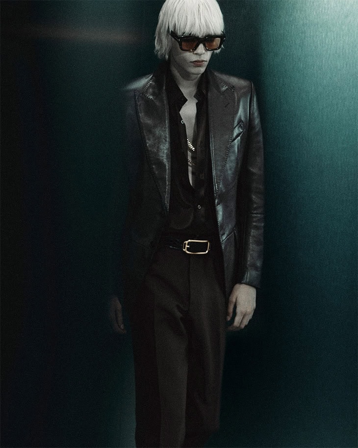 Adult male with blond hair poses in black leather jacket, dark collared shirt, and dark trousers in a dark indoor environment.