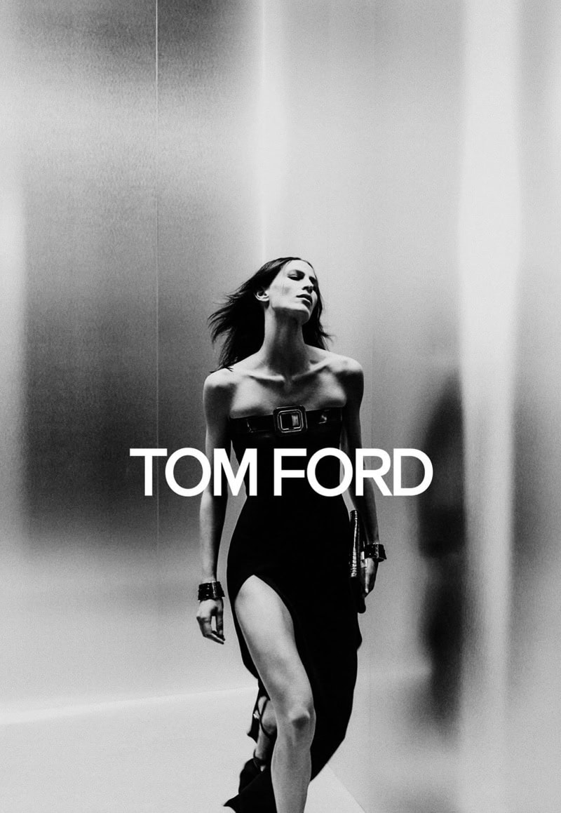 Black-and-white fashion portrait of a slender woman in a strapless dress with a thigh-high slit, posing against a blurred studio backdrop.