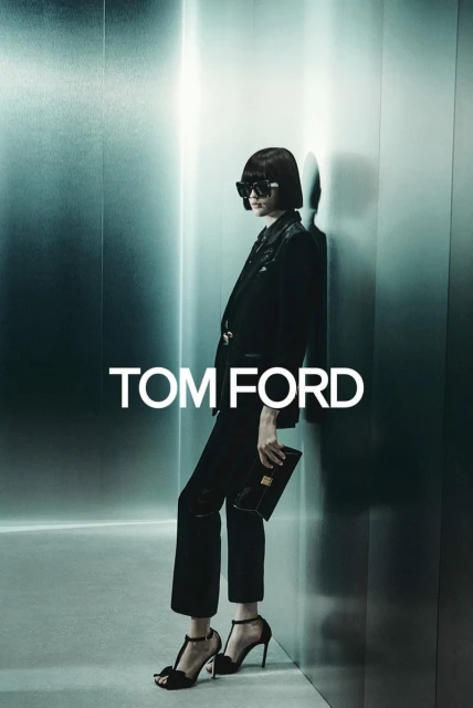 Fashion shot: Woman in tailored black suit, black heels, sunglasses, clutch, posing in a photo studio against a textured gray wall.