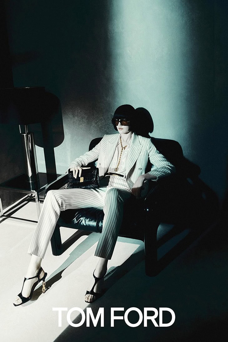 A woman sits in a leather chair wearing a striped suit, accessorized with dark glasses, a necklace, a bag, and strappy high heels.