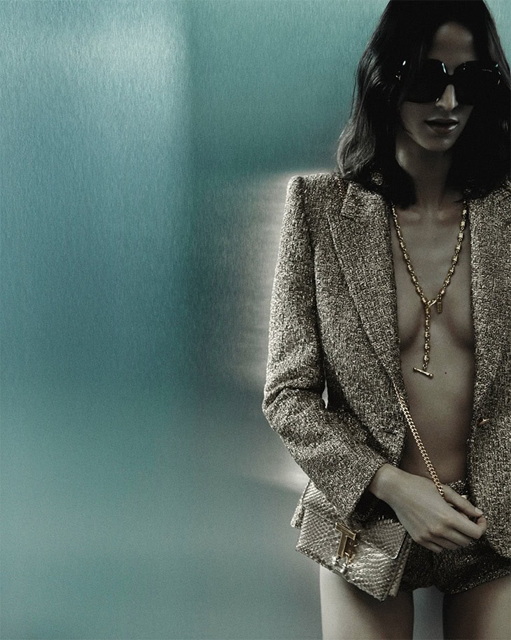 Adult woman wearing a gold jacket, shorts and sunglasses with a gold chain, posing against a blue metallic background in a studio shot.