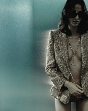Adult woman wearing a gold jacket, shorts and sunglasses with a gold chain, posing against a blue metallic background in a studio shot.