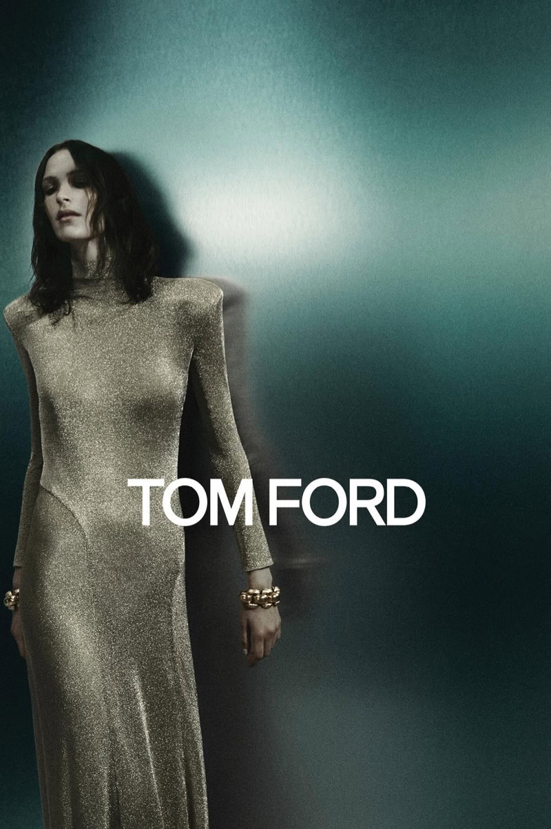 Woman in gold, long-sleeved gown, accessorized with bracelets, stands against a textured blue background with brand name overlay, "Tom Ford."