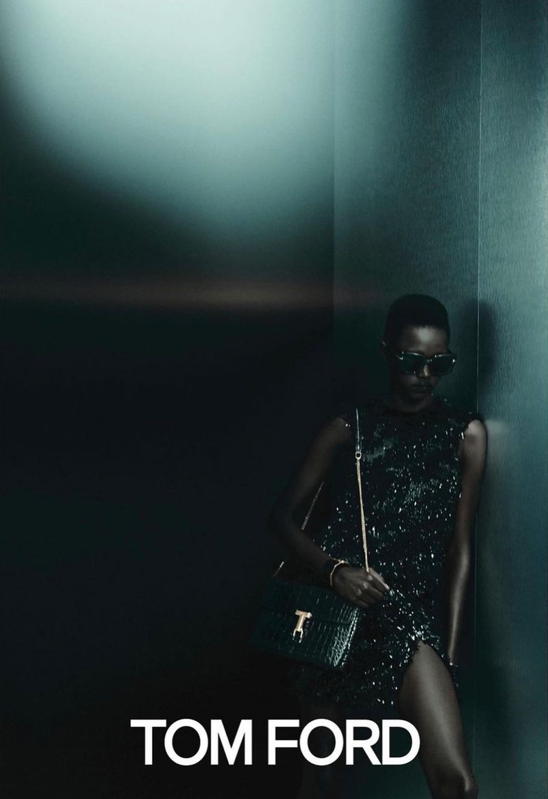 Fashionable woman in dark sunglasses and a glittering dress poses with a green handbag in a dimly lit interior, promoting Tom Ford.