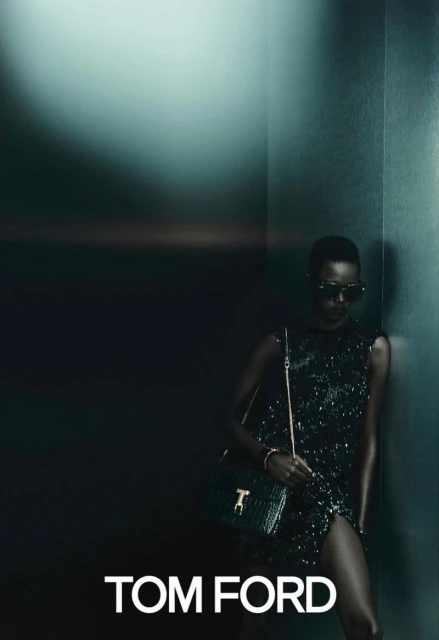 Fashionable woman in dark sunglasses and a glittering dress poses with a green handbag in a dimly lit interior, promoting Tom Ford.