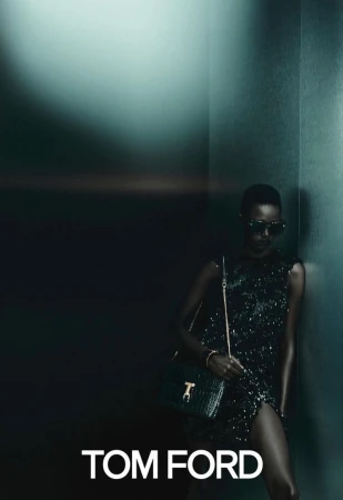 Fashionable woman in dark sunglasses and a glittering dress poses with a green handbag in a dimly lit interior, promoting Tom Ford.