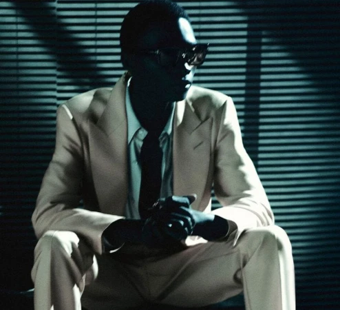 Fashionable person wearing a light suit and sunglasses seated in front of blinds, with hands clasped, set against a dim and stylish backdrop.